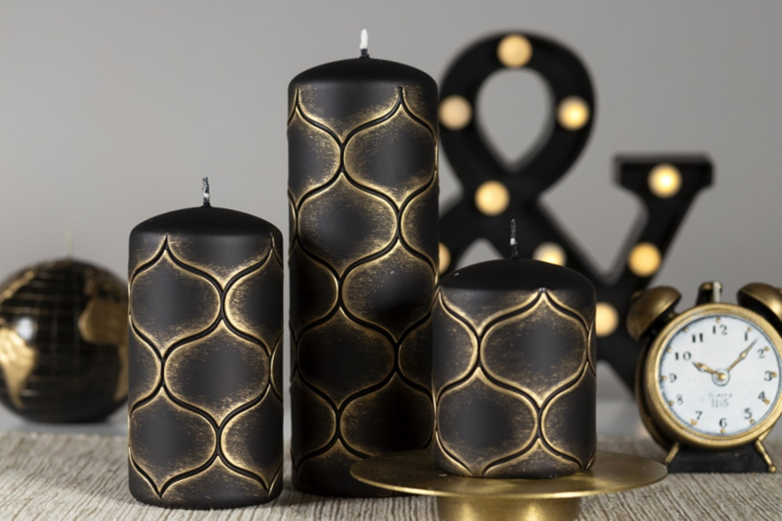 Three pillar candles with golden Moroccan pattern beside vintage alarm clock and globe