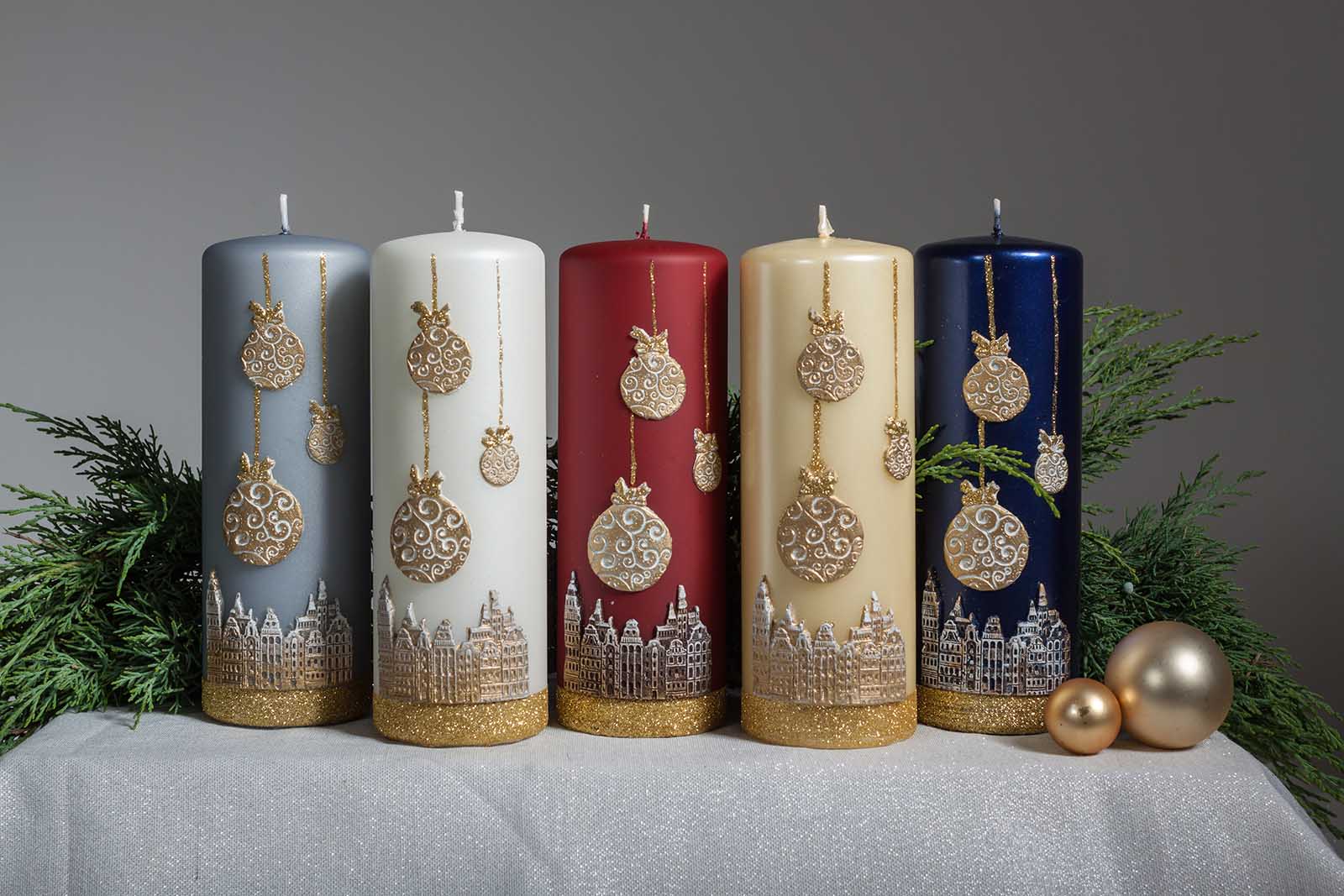 Five Christmas themed candles sitting beside two Christmas bubbles and with pine leaves at the background