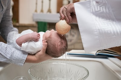 Father is holding baby over the crystal bowl while priest is pouring holy water on the child’s head