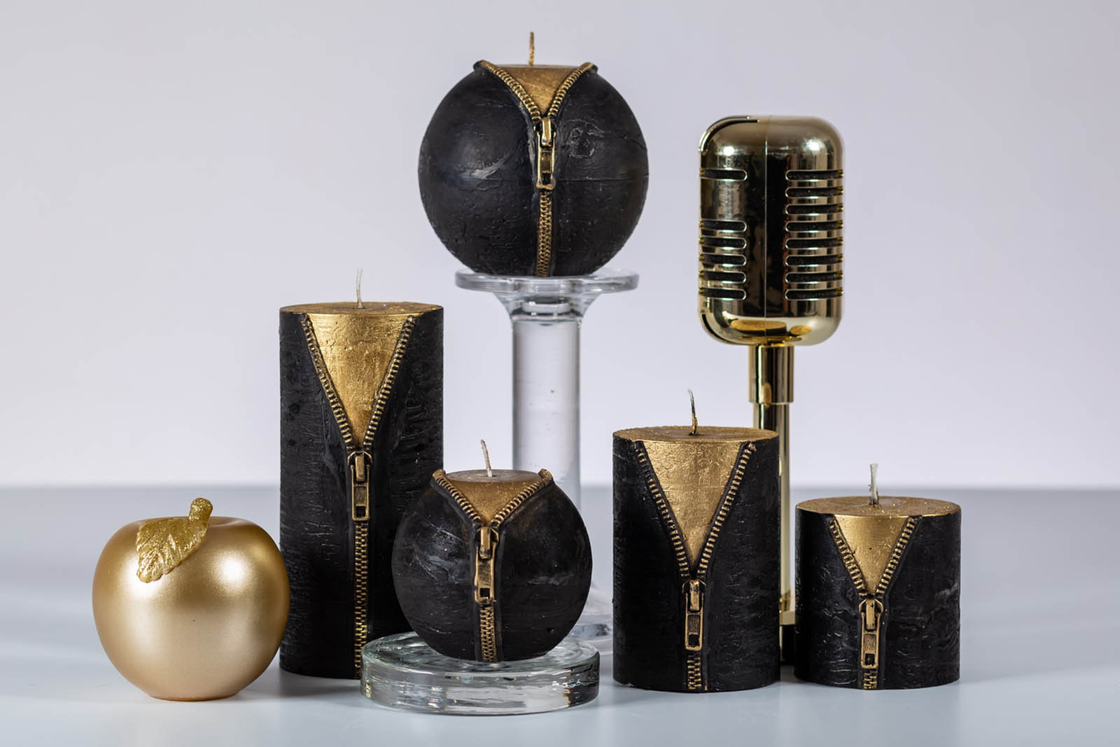 Five black and gold rustic candles with zipper sitting on the beige tabletop beside golden microphone and apple