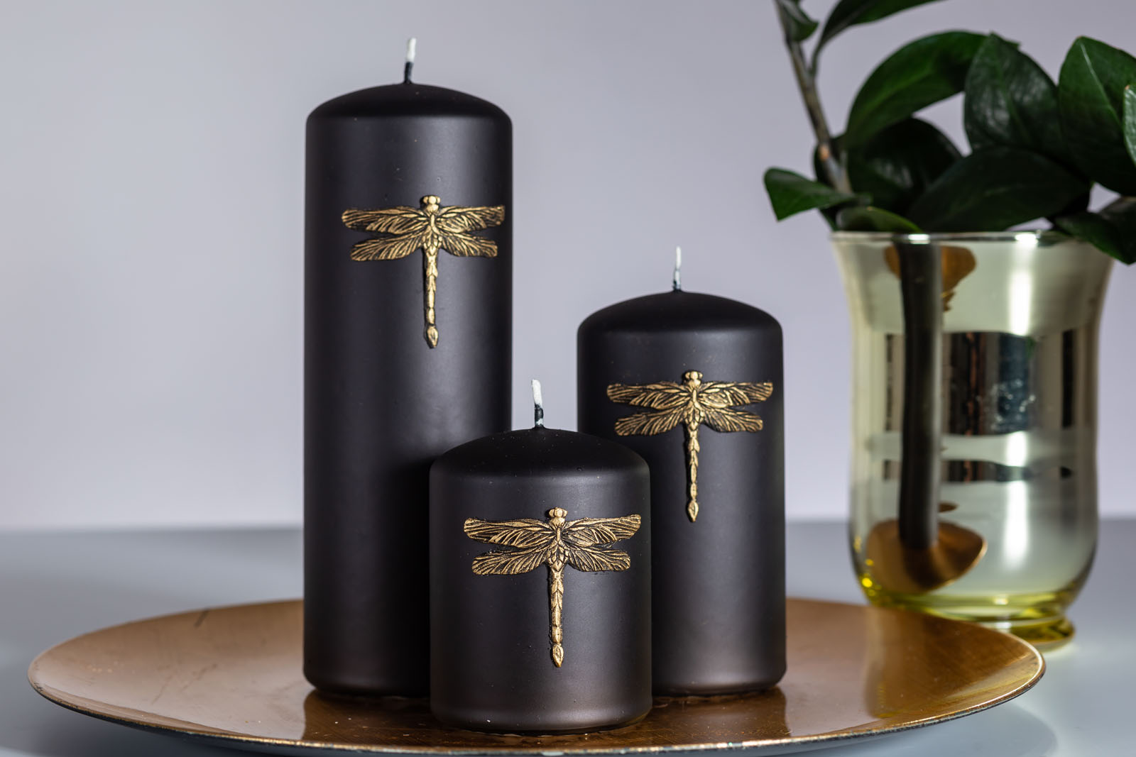 Three pillar candles with golden dragonfly sitting on the brown plate beside flower vase with flowers